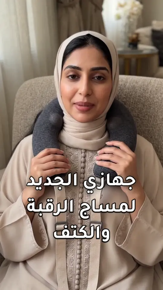 A frame from the generated video ad: a Gulf woman in a beige abaya on a sofa, the neck massager resting on her shoulders, with the burned-in Arabic caption “جهازي الجديد لمساج الرقبة والكتف”.